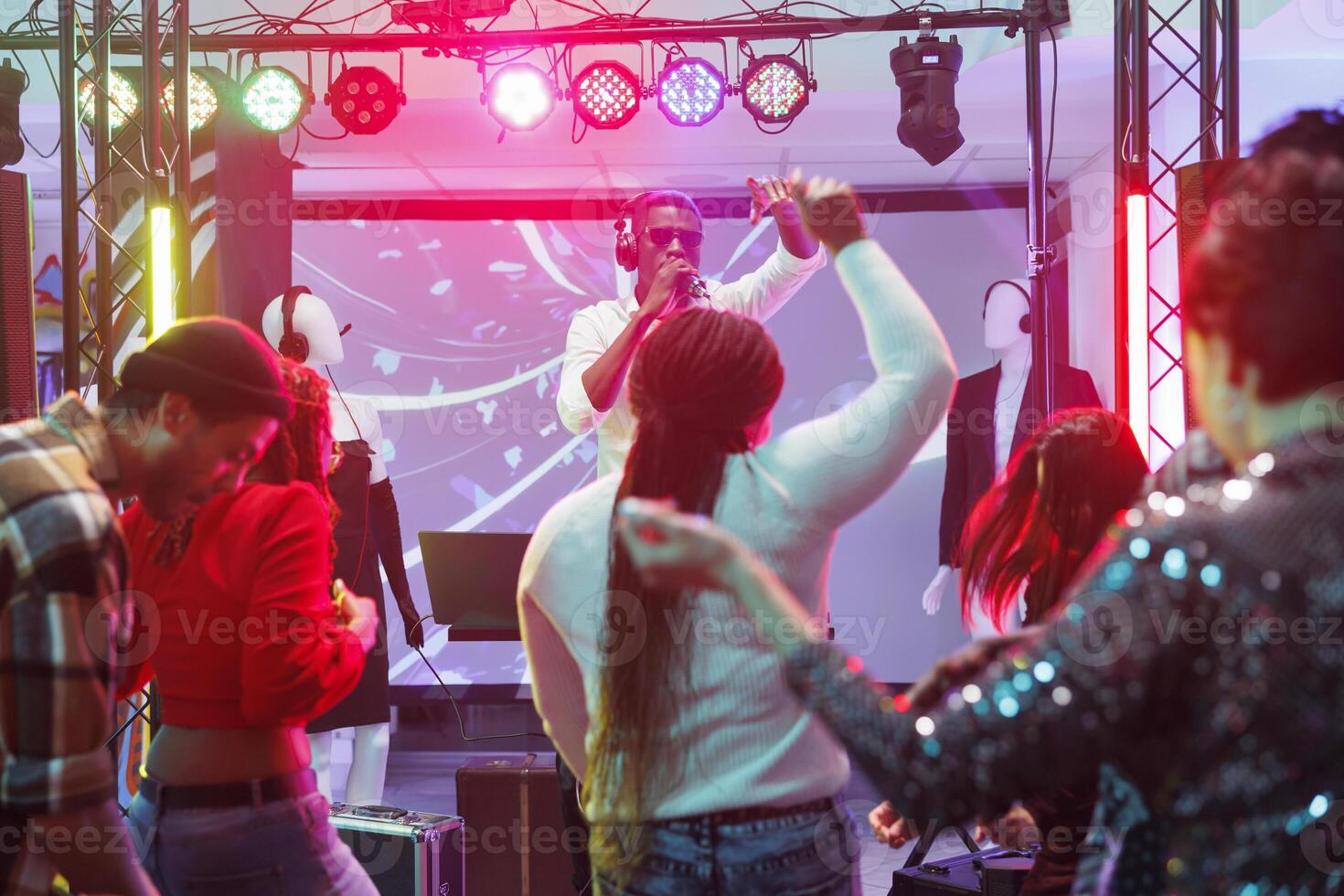 Dj speaking to audience in microphone while performing on stage in nightclub. People dancing and raising hands on dancefloor while attending electronic music concert in club photo