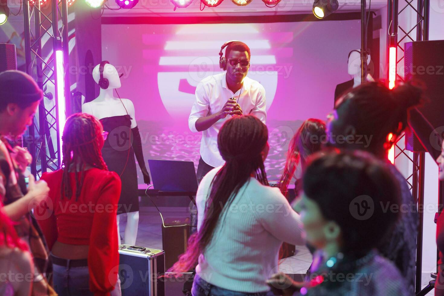 Dj mixing music and singing in microphone while crowd dancing and cheering in nightclub. African american man musician performing on stage and speaking to audience in club photo