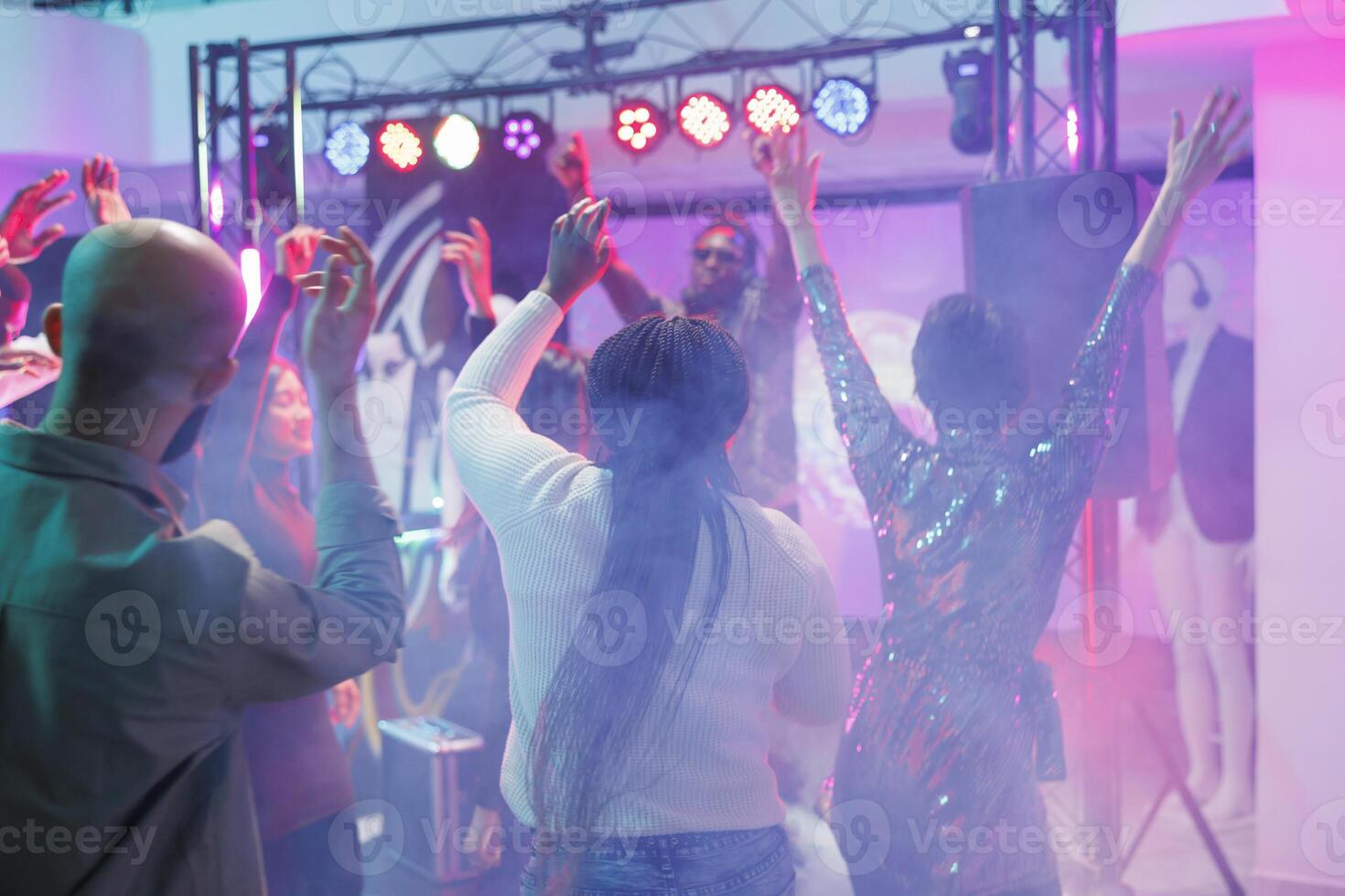 Energetic crowd dancing and enjoying discotheque on dancefloor with colorful spotlights. People jumping, having fun and moving to music beats with raised hands in nightclub photo