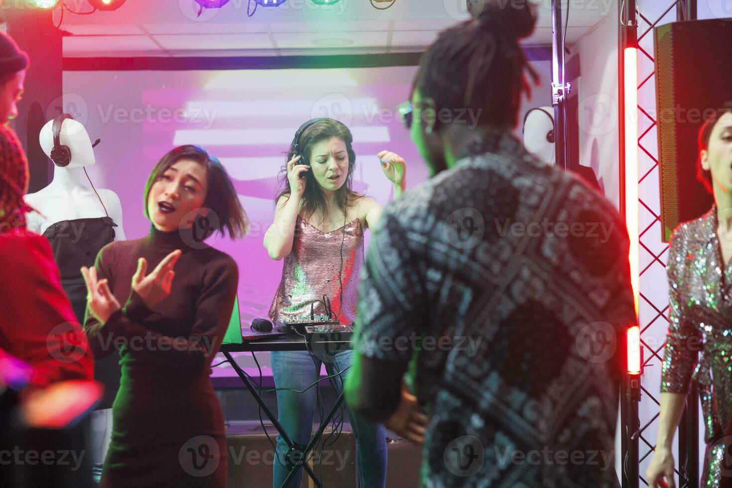 Woman dj in headphones performing using electronic music mixing station on stage while crowd dancing in nightclub. People partying on dancefloor and clubbing at discotheque photo