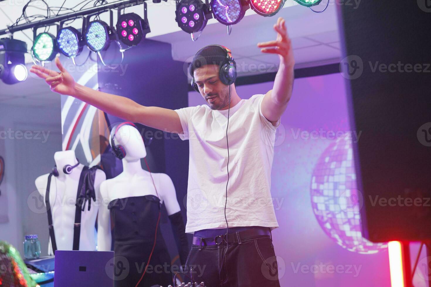 Young dj in headphones performing on stage and raising hands while mixing electronic music in club. Man musician wearing earphones playing in nightclub at discotheque party photo
