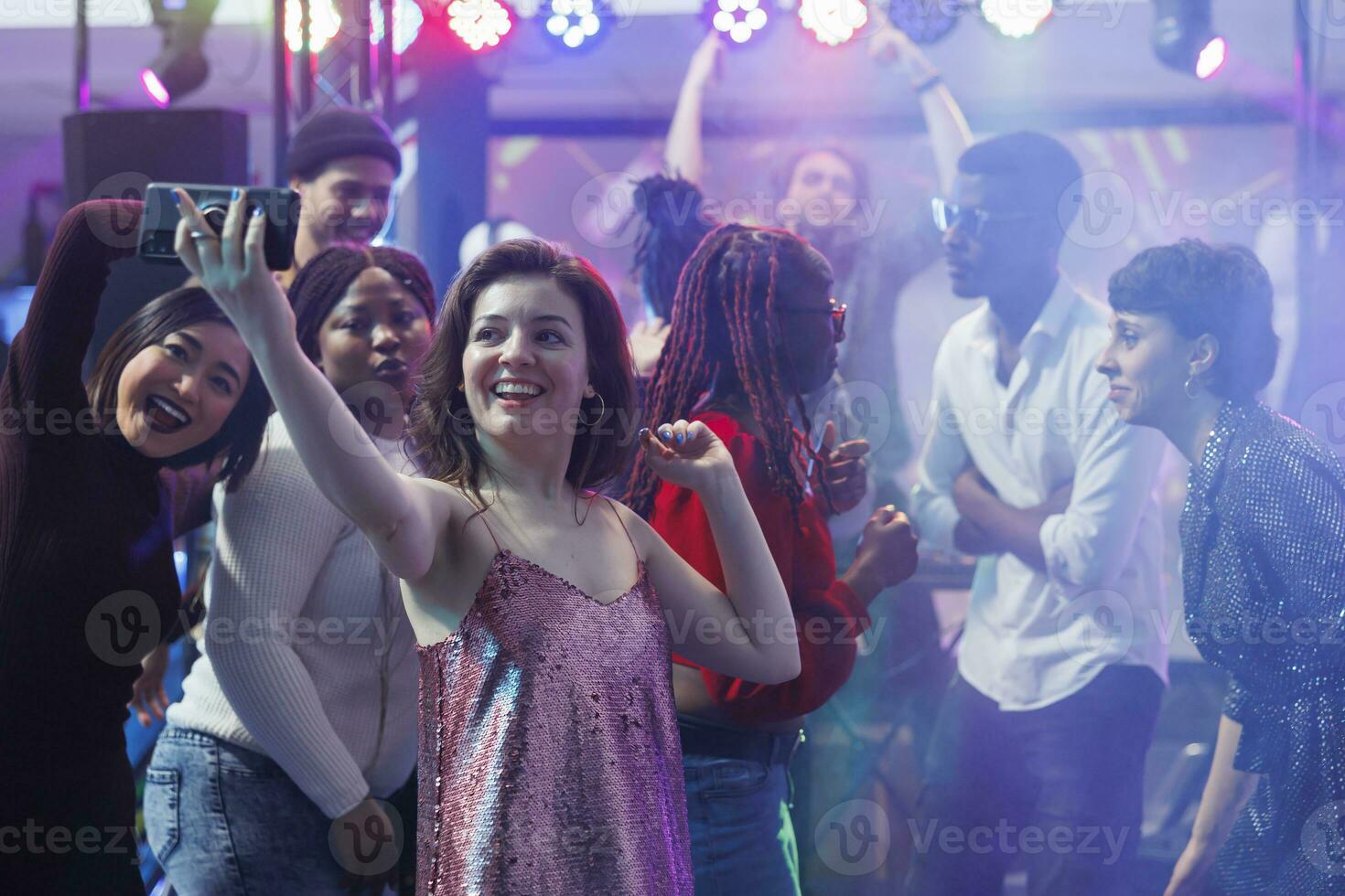 Smiling woman taking selfie while dancing with friends at nightclub discotheque. Happy girl holding mobile phone and posing for photo on club dancefloor illuminated with stage spotlights