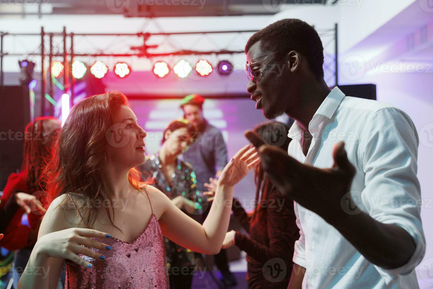 Man and woman diverse friends dancing together and chatting at nightclub disco party. Young multiracial couple clubbing and having fun on dancefloor at discotheque in club photo