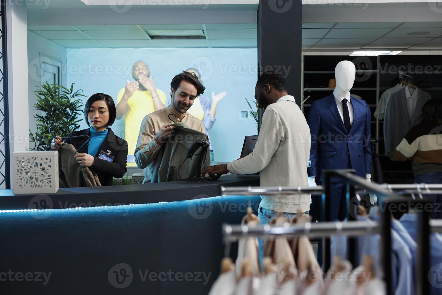 Smiling customer and cashier chatting at checkout while purchasing clothes in shopping mall fashion department. Store employee scanning garment and chatting with african american client photo