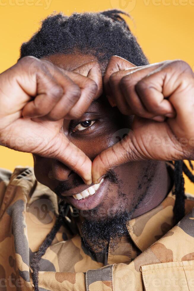 Lovely person doing heart shape in front of camera, smiling during valentine s day studio shot. African american man making love symbol with fingers over yellow background. Romantic gesture photo