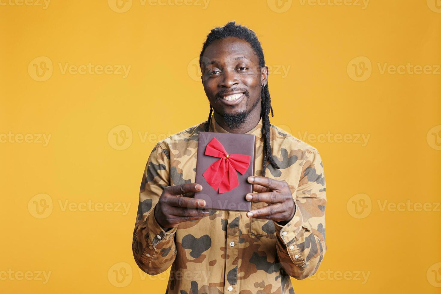 Lovely african american man showing wrapped gift box, presenting girlfriend present for valentine s day in studio over yellow background. Cheerful smiling person celebrating love day photo