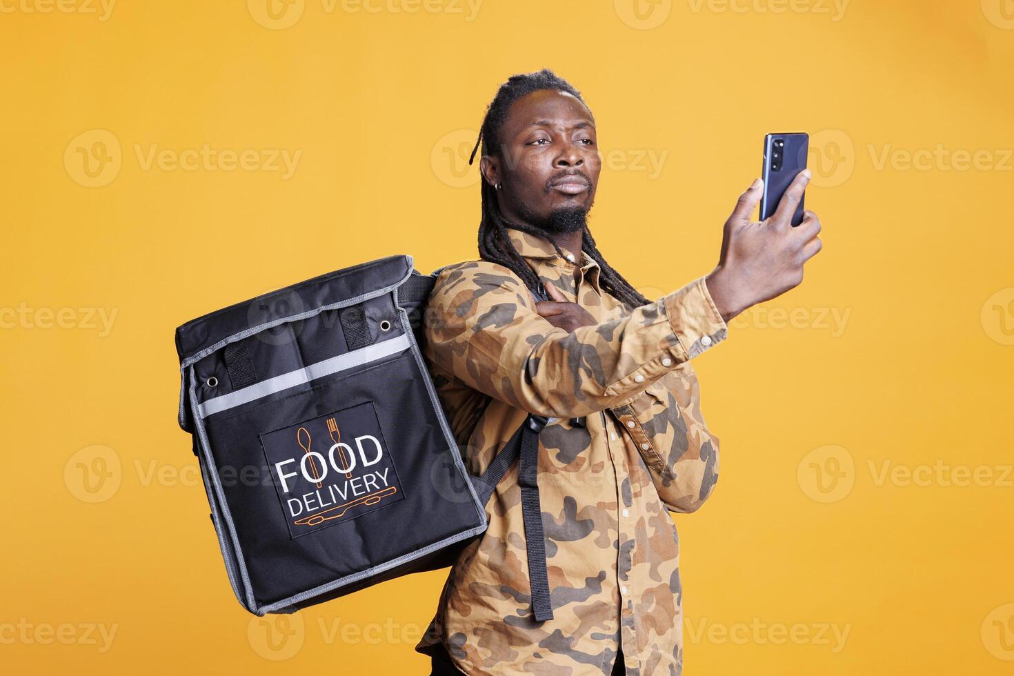 Deliveryman holding phone checking client adreess using takeaway food app before delivery food order. African american restaurant worker carrying thermal backpack standing in studio photo