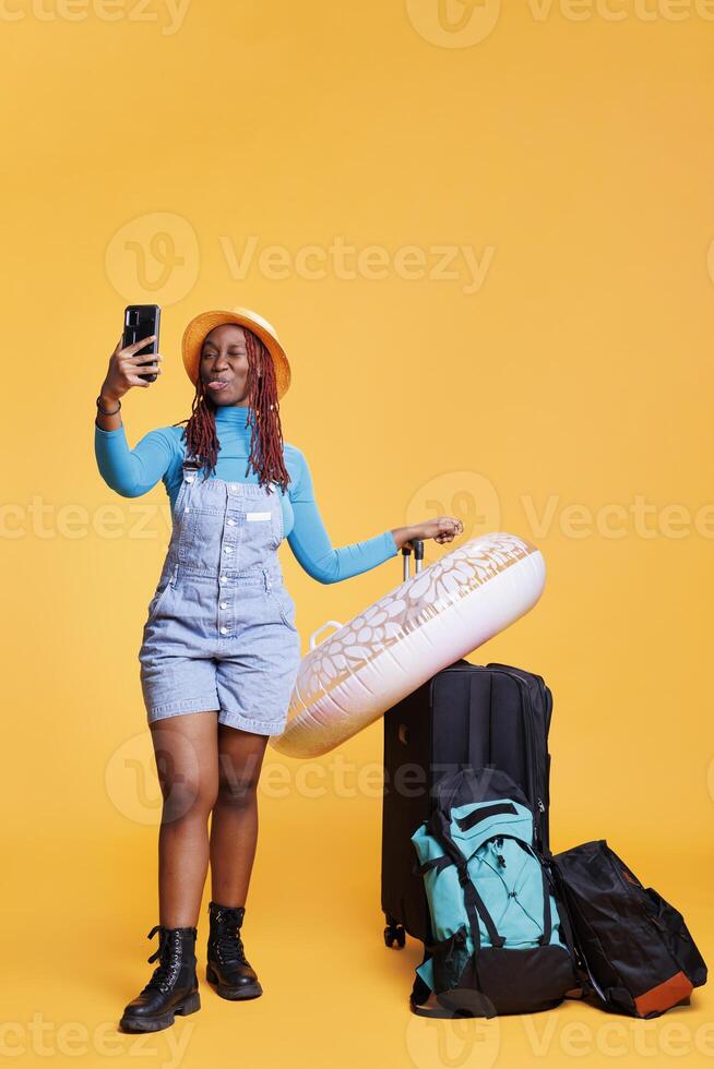 joven adulto tomando imágenes en viaje, que lleva equipaje carretilla pantalones y inflable en cámara. alegre viajero teniendo divertido con fotos en teléfono inteligente aplicación, fin de semana urbano aventura.