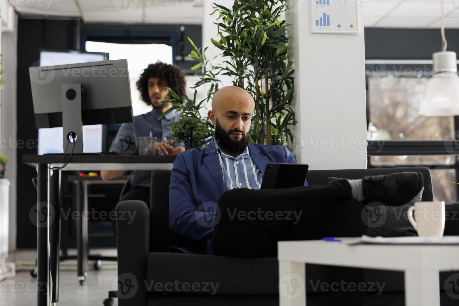 Arabian businessman analyzing financial data on digital tablet in coworking space. Executive manager sitting on couch while working on business development project in corporate office photo