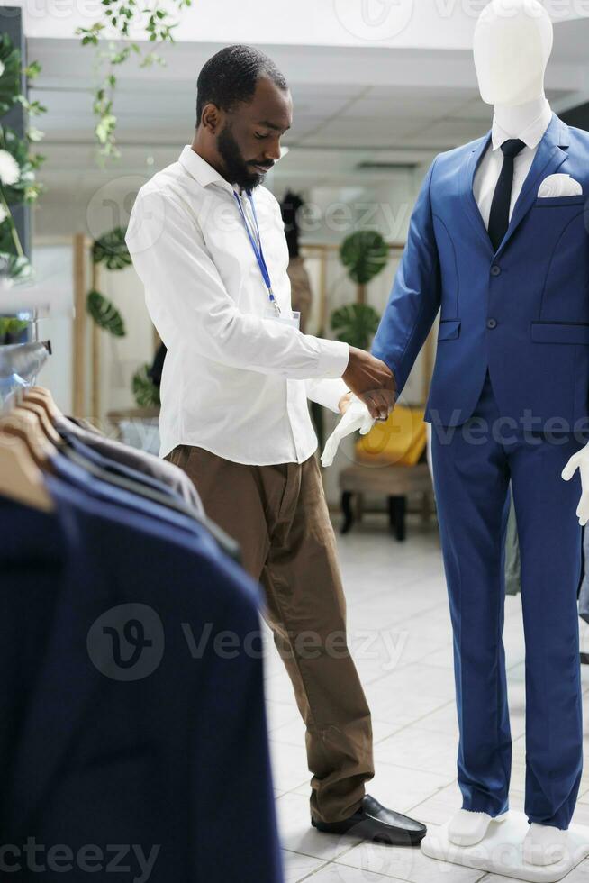 Assistant adjusting jacket on mannequin wearing formal outfit in clothing store. African american man checking male suit on model showcasing apparel for customers in shopping mall photo