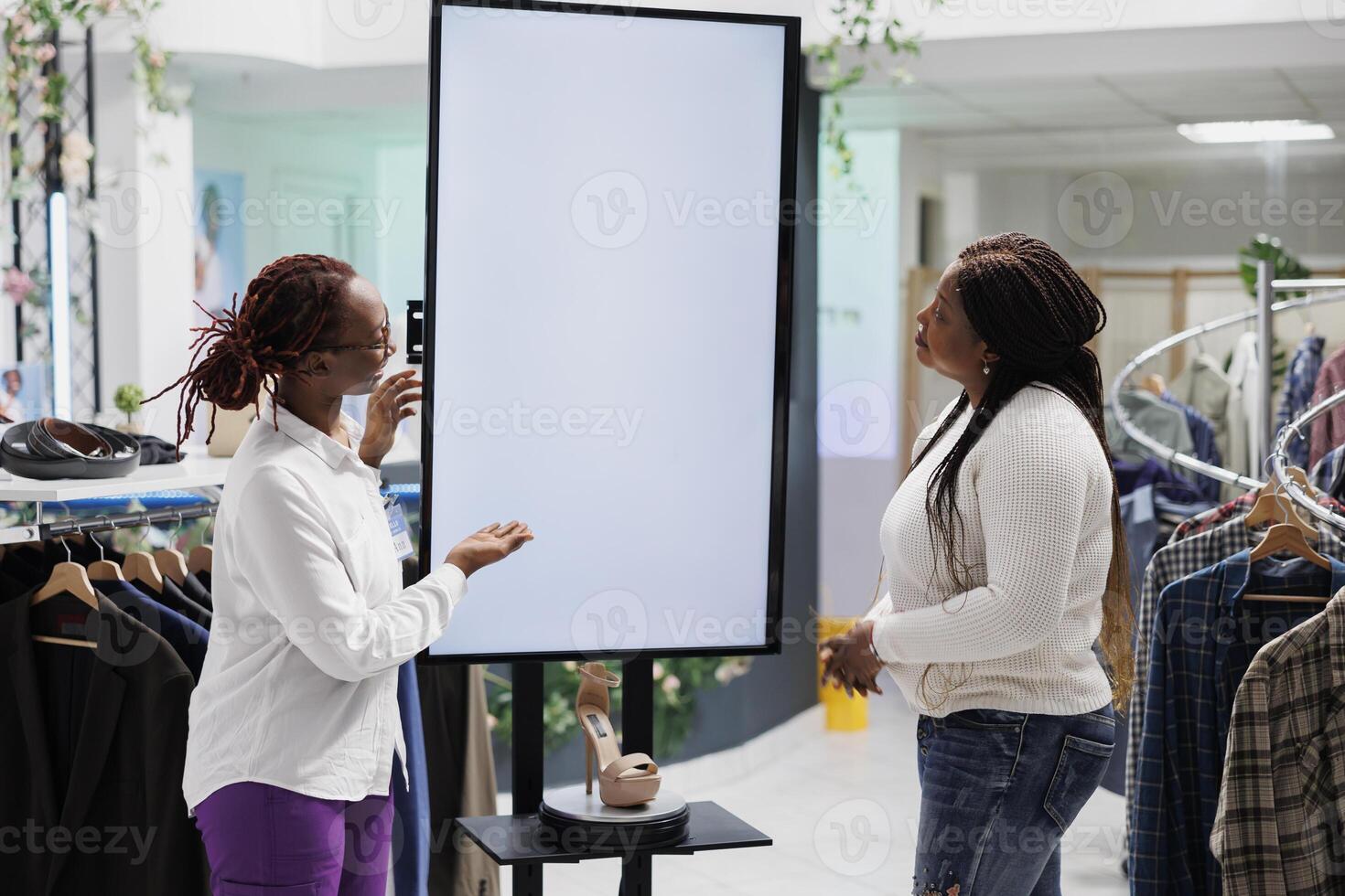 Clothing store worker helping woman to choose shoes, showing options on empty whiteboard mock up. Mall assitant advertising stiletto to buyer using promotion on digital blank smart screen photo