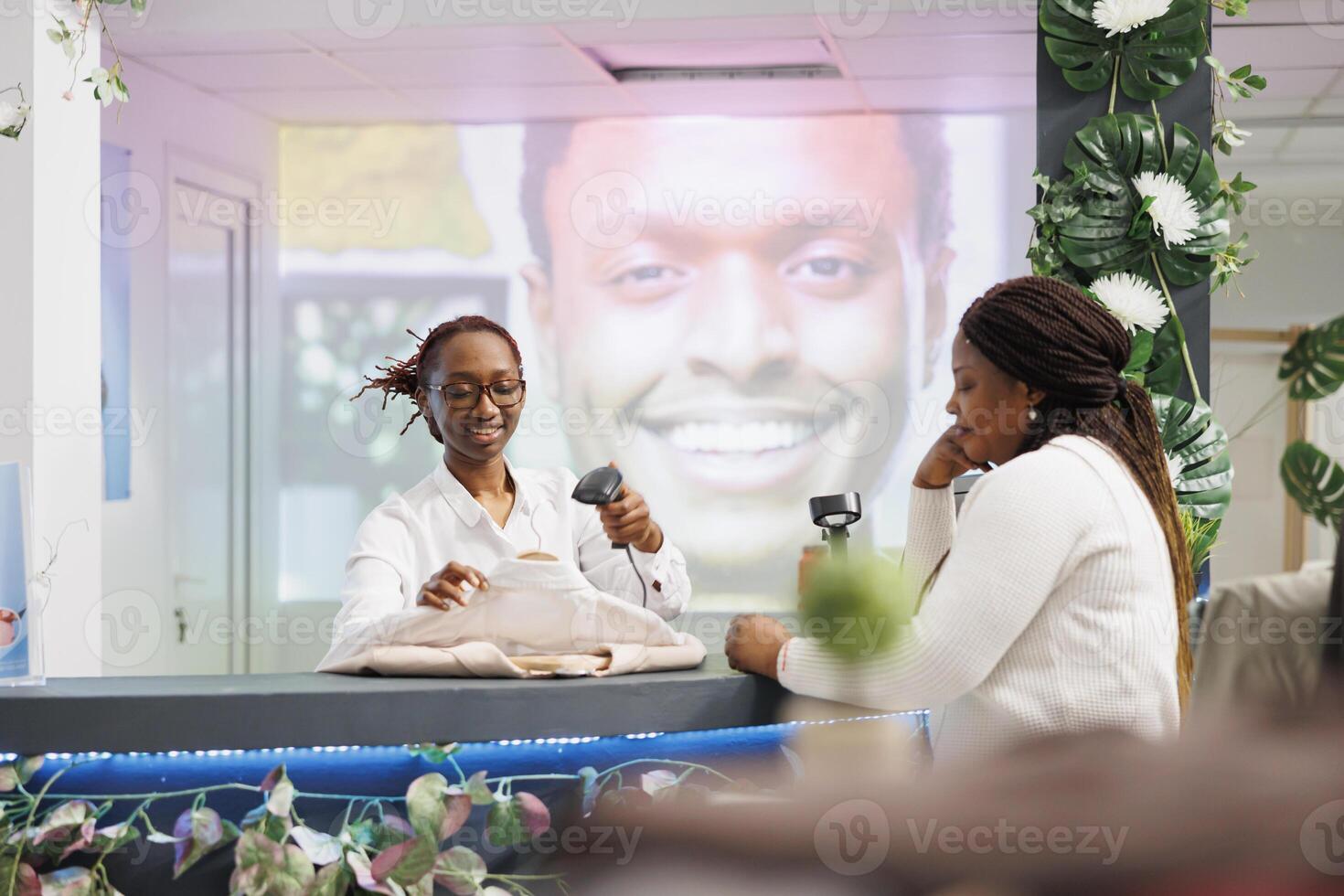 Smiling african american cashier scanning customer clothes at cash register. Woman standing at counter desk, waiting to pay for purchase while employee using pos terminal and scanner photo