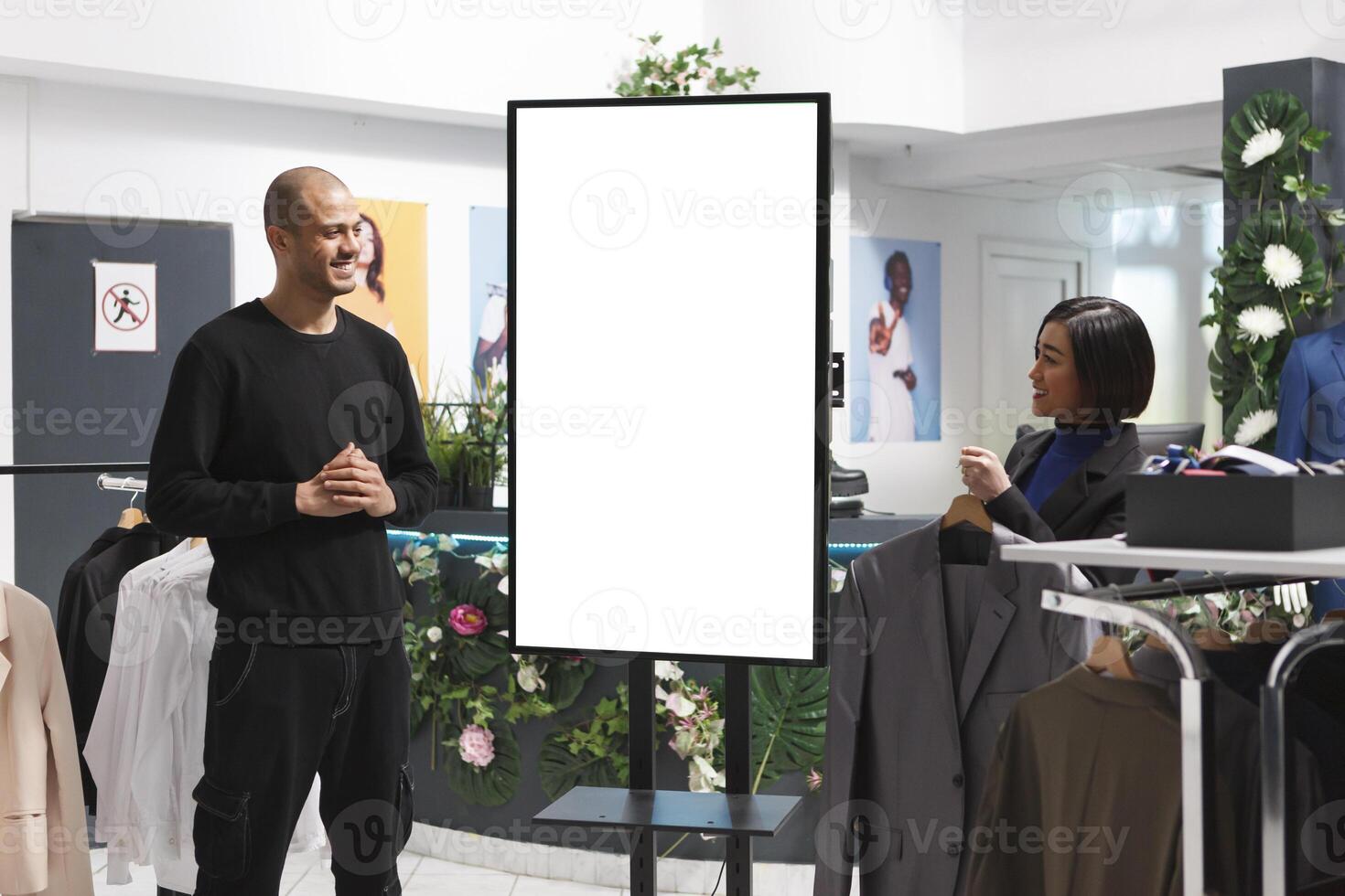 Smiling clothing store assistant chatting with arab customer while standing near blank advertising whiteboard. Shopping center asian woman employee helping client to choose apparel photo