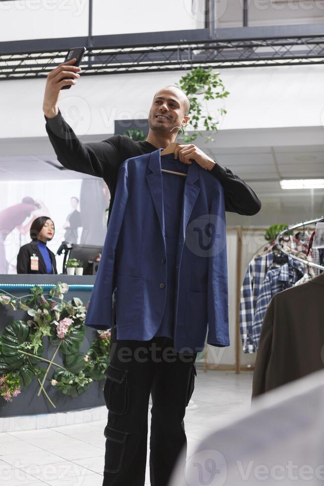 ropa Tienda árabe hombre cliente participación chaqueta en percha y tomando foto en teléfono inteligente mientras hojeada vestir en compras centro comercial. boutique sonriente comprador chateando en videollamada y demostración atuendo