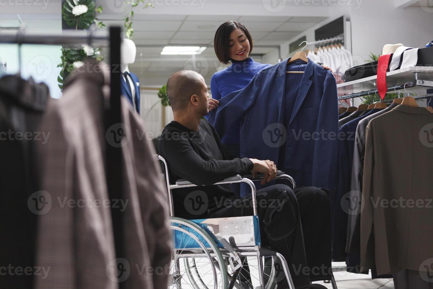 ropa Tienda sonriente asistente Ayudar cliente en silla de ruedas a examinar chaqueta estilo y adaptar. compras centro comercial asiático mujer vendedor consultante árabe hombre con invalidez y dando Moda Consejo foto