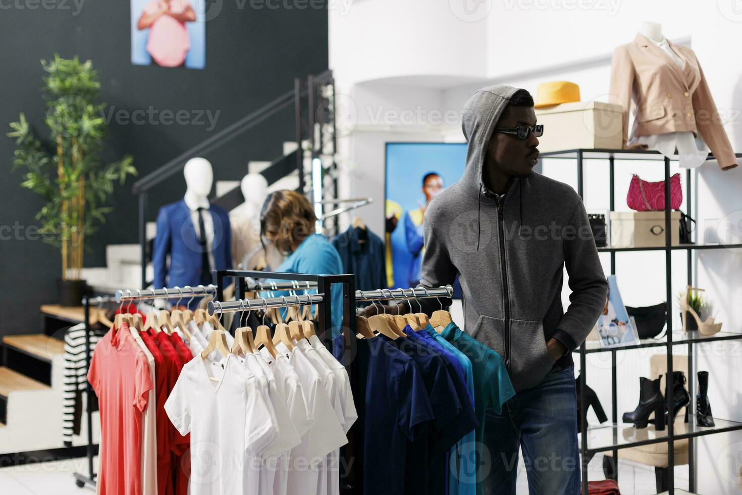 African american man with hood and glasses, trying to steal fashionable merchandise from clothing store. Robber looking around to see if someone watching him while stealing trendy clothes photo