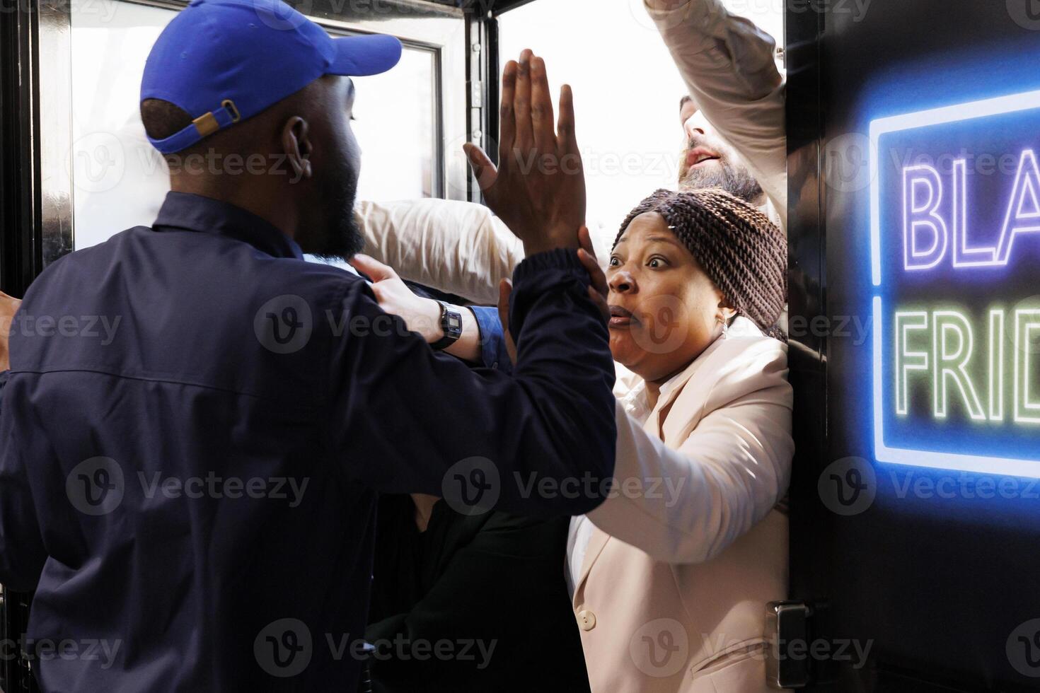 Rude aggressive shoppers try to enter into shopping mall and get best Black Friday deals first. Store security officer holding back angry crazy crowd of diverse people breaking into store during sales photo