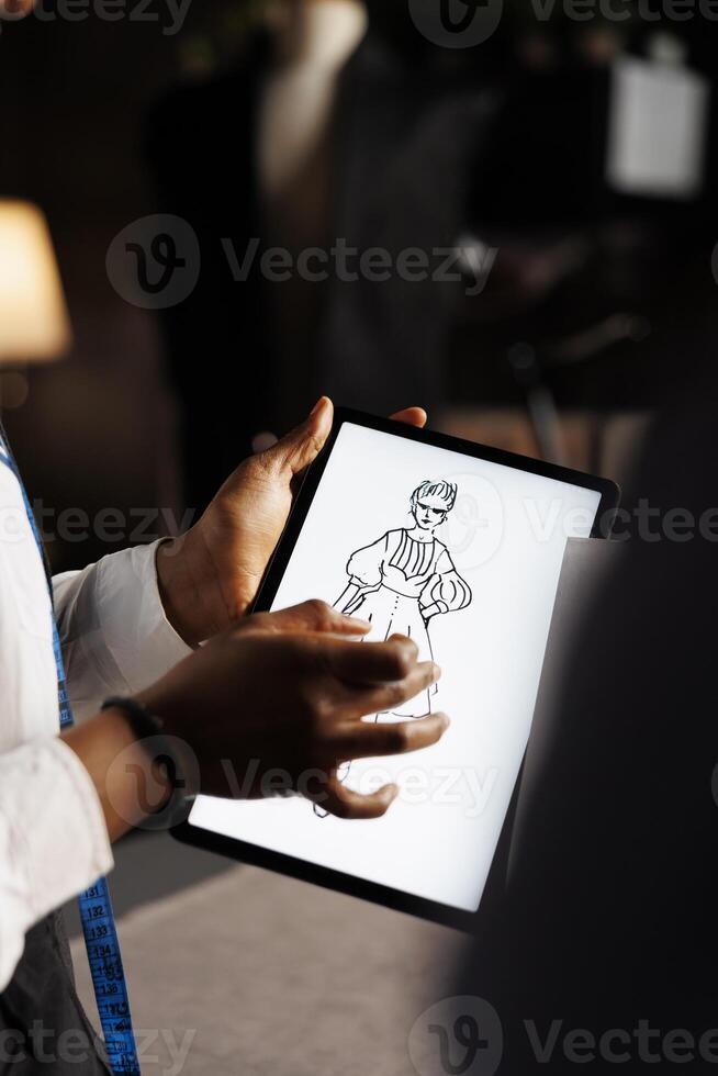 African american couturier using sketch drawing on tablet to measure fit adjustments to be made in tailoring studio. Fashion designer working on stylish bespoke sartorial dress photo