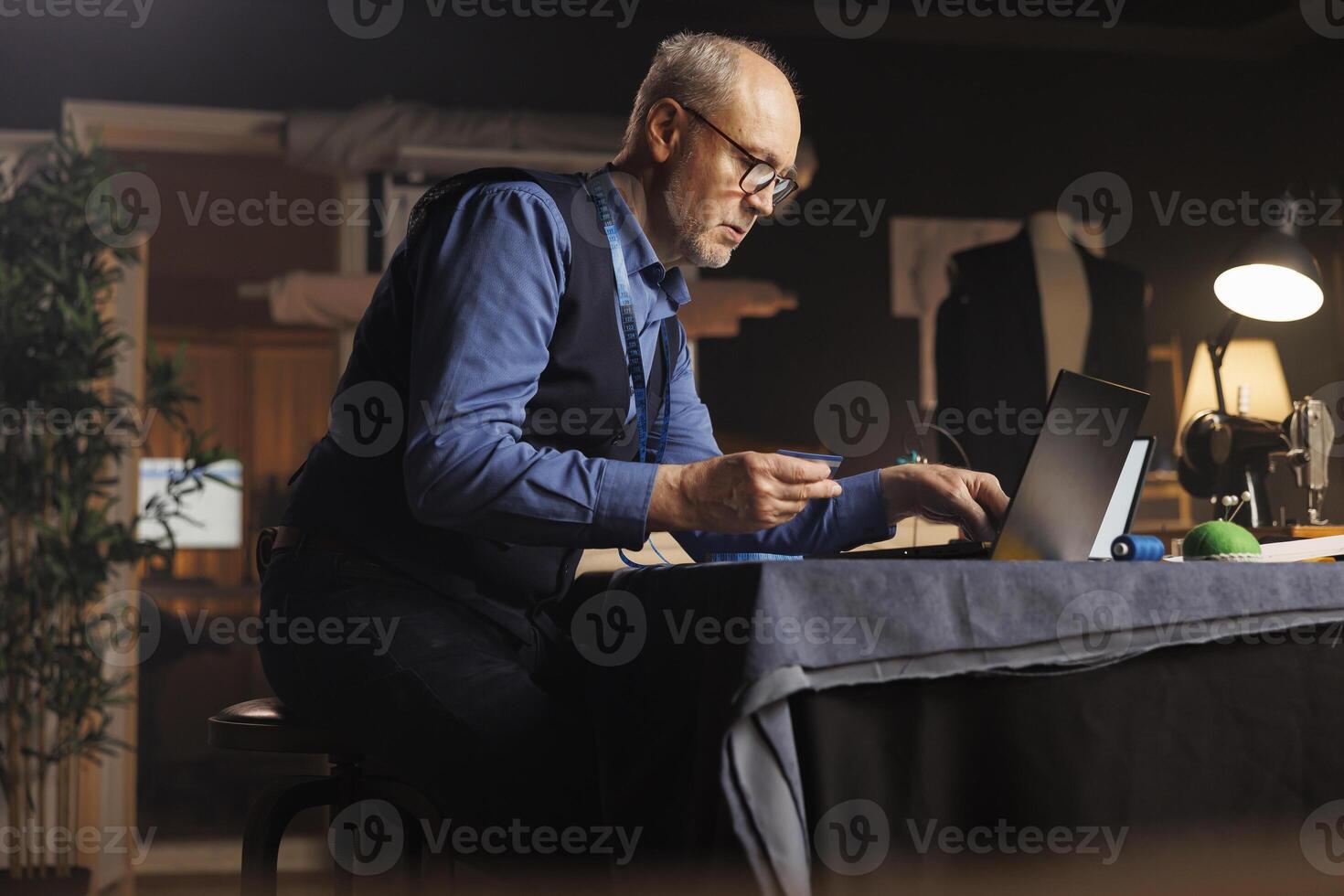 Fashion designer buying sewing materials online for upcoming customer comission tailoring modifications. Elderly couturier imputing credit card purchasing details on laptop in atelier studio photo