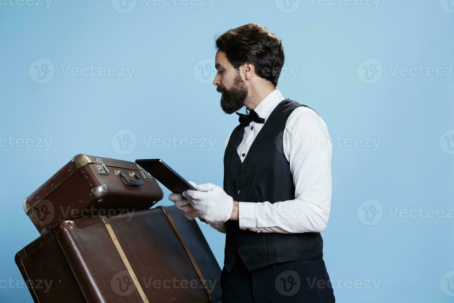 Hotel concierge counting trolley bags and checking tablet with list of reservations, studio shot with blue background. Young professional doorman with gloves taking care of luggage. photo