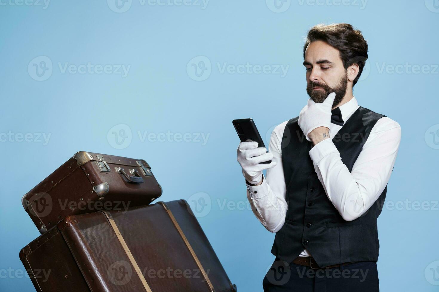 While behaving as skilled member of elegant hotel staff on camera, bellboy uses smartphone to access website. Reservation inquiries get approved electronically by the receptionist. photo