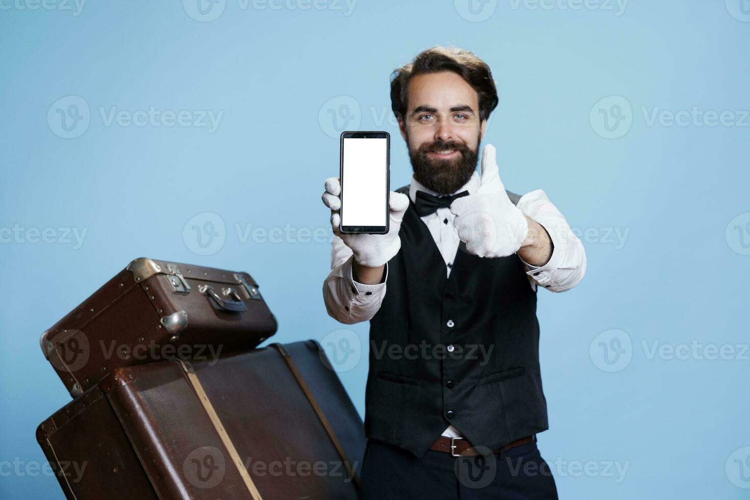 Classy hotel concierge next to luggage holding phone with white empty display, tourism concept. Bellboy showing blank screen on camera, commitment of his professional duties. photo