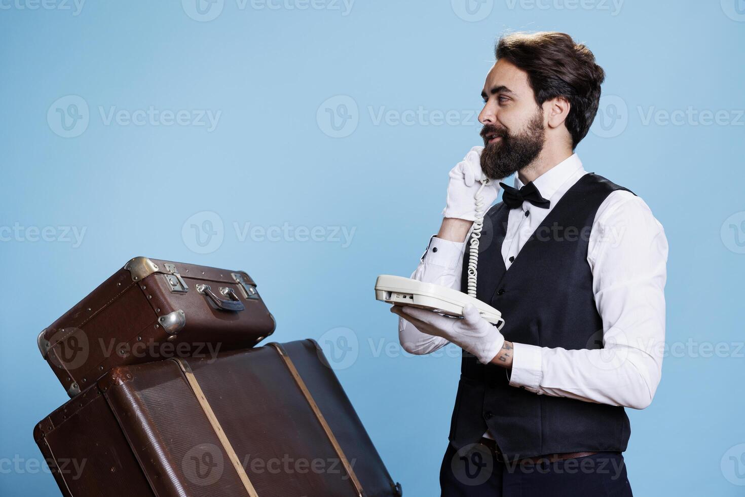 Elegant bellboy chats with guests about reservations as they call on hotel landline phone. Classy hotel concierge using telephone with cord to talk about services, professional industry. photo