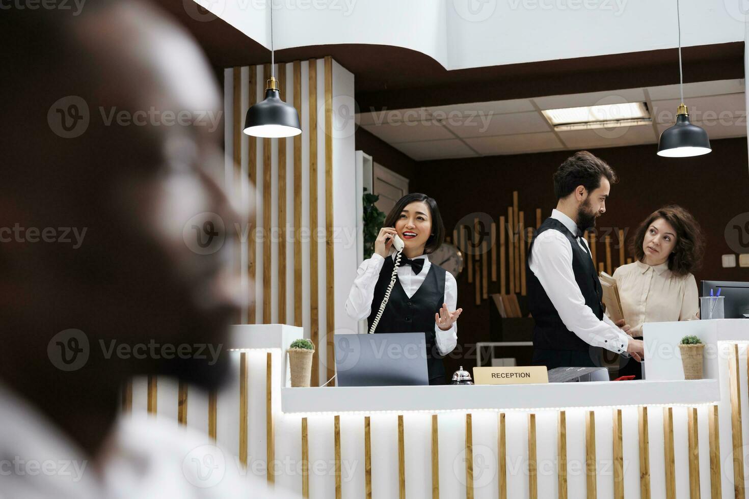 Diverse people standing at front desk working on room reservations, answering landline phone call to note booking on hotel record files. Reception staff giving luxury service for guests. photo