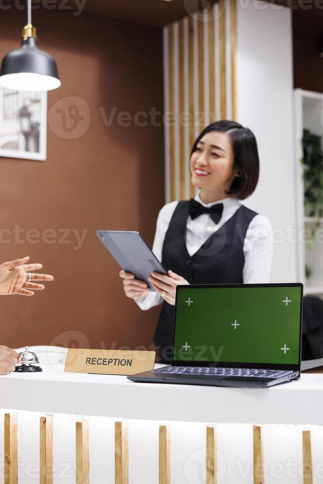 Receptionist having greenscreen on desk, welcoming guests at hotel and starting check in process with tablet. Asian woman using laptop showing isolated display with chroma key copyspace. photo