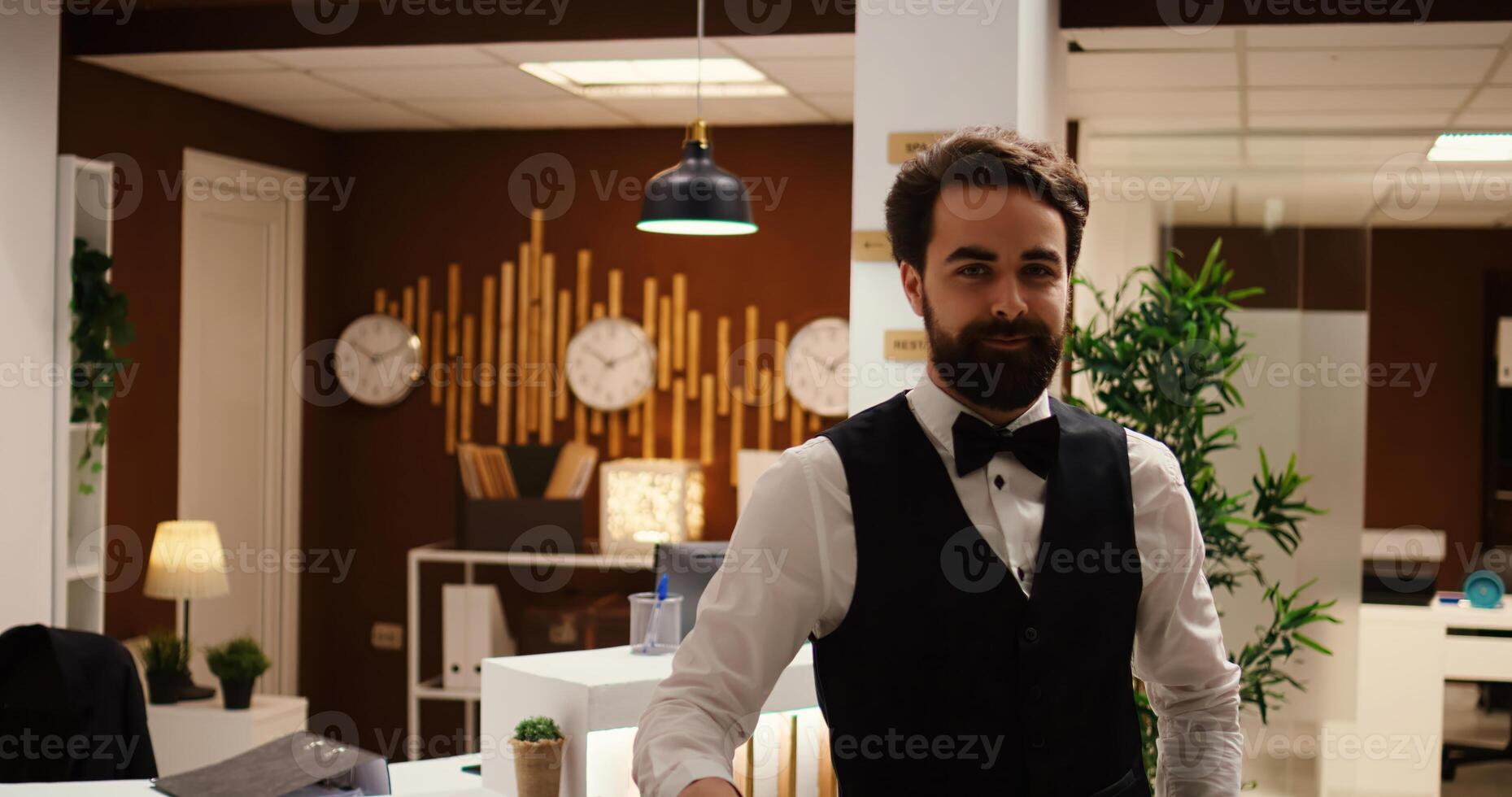 Happy elegant professional hotel staff worker standing at check in reception desk, close up. Portrait of cheerful smiling bellhop employee standing by baggage trolley in travel resort lobby photo