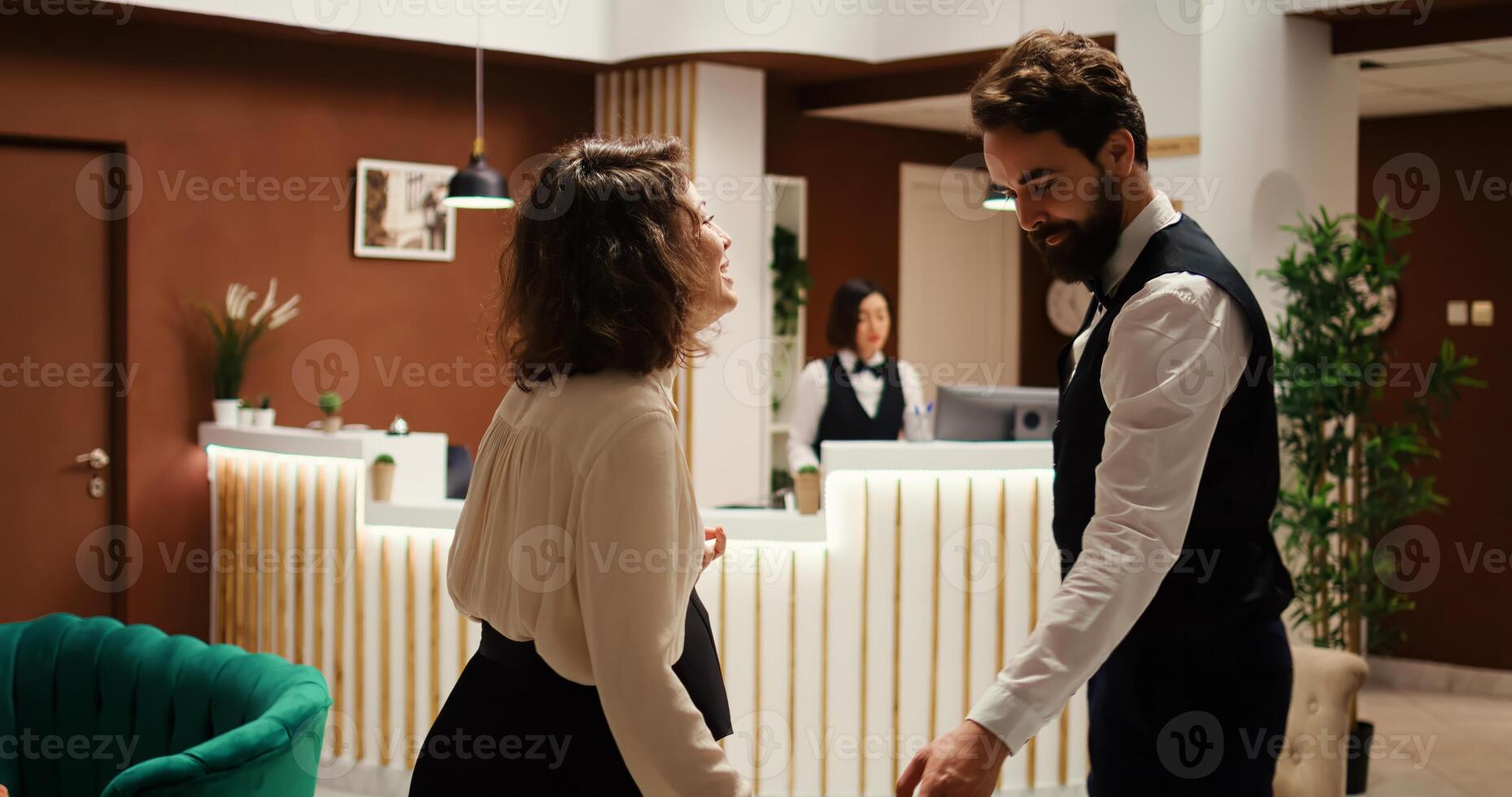 Businesswoman checking in luxury hotel, being helped by bellboy with luggage while receptionist checks booking details. Resort visitor in professional trip assisted by concierge personnel photo