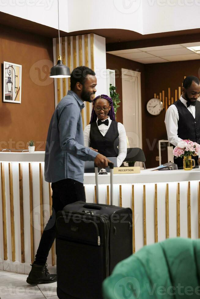 Happy young African American man arriving at resort, standing with suitcase at reception area checking in into hotel, enjoying excellent customer service. Smiling black guy tourist at front desk photo