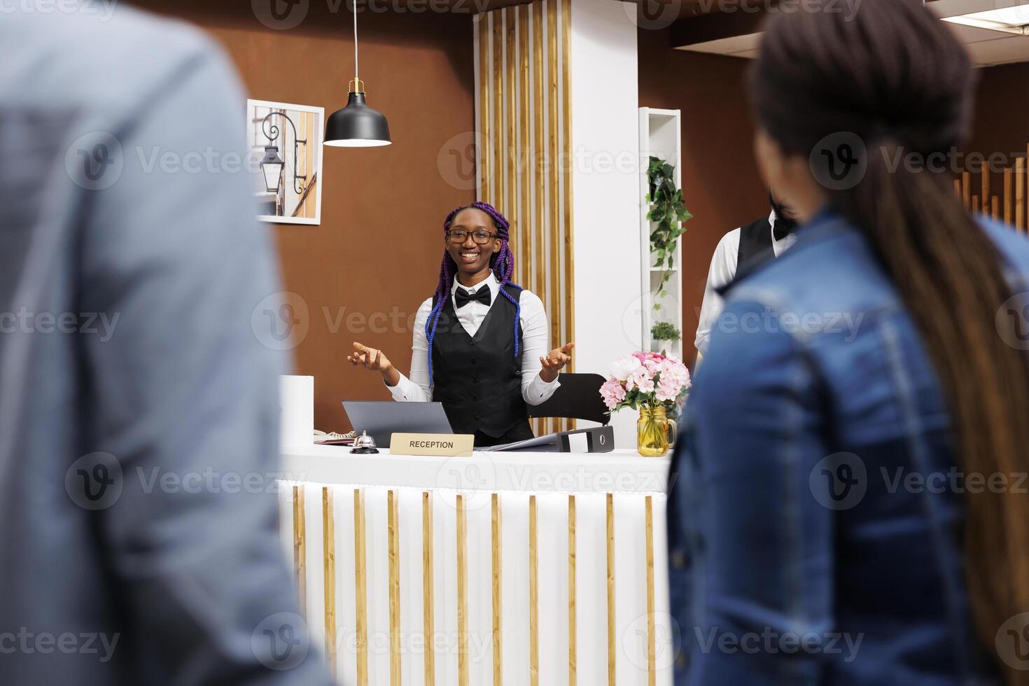 Friendly young African American female receptionist standing at hotel reception greeting tourists. Positive black girl front desk agent wearing uniform smiling to arriving guests photo