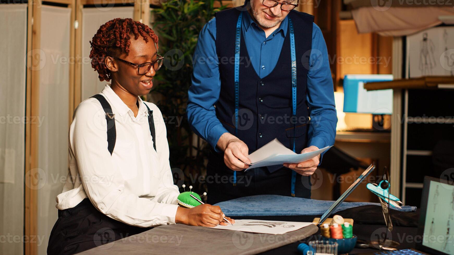 Tailor and apprentice reviewing sketches in atelier, trainee learning handcraft concept to create clothes. Experienced couturier explaining needlework skills to beginner in tailor workshop. photo