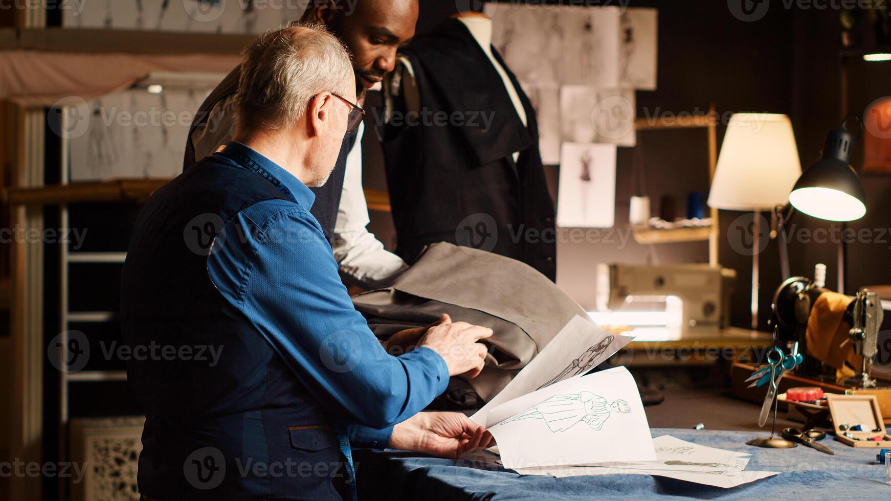 Old craftsman and student making sketch to design new fashion items, apprentice learning about clothes manufacturing and textile industry. Experienced tailor teaching trainee. Tripod shot. photo