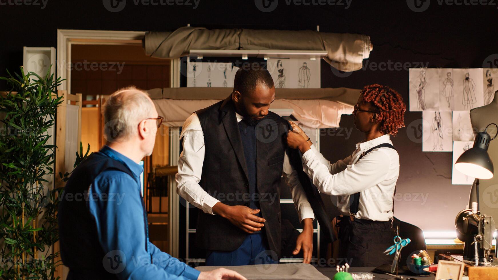 Young customer trying on suit jacket in atelier, making sure it fits with the right measurements. Experienced tailor and assistant designing custom made blazer, fitting in workshop. photo