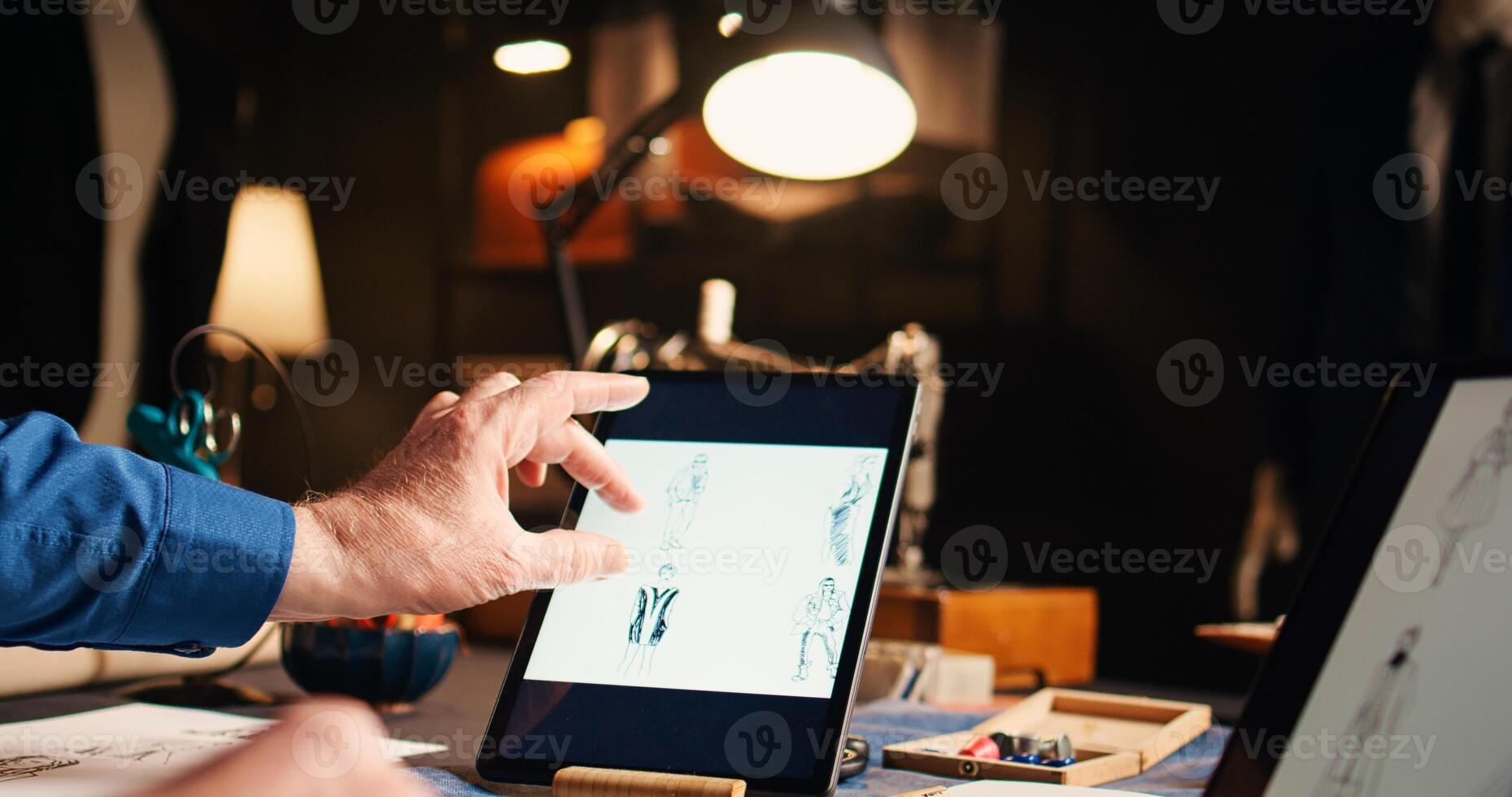 Old couturier checking fashion sketches in atelier, working on modern designs with laptop and tablet. Experienced designer searching handmade models online, needlework. Handheld shot. photo