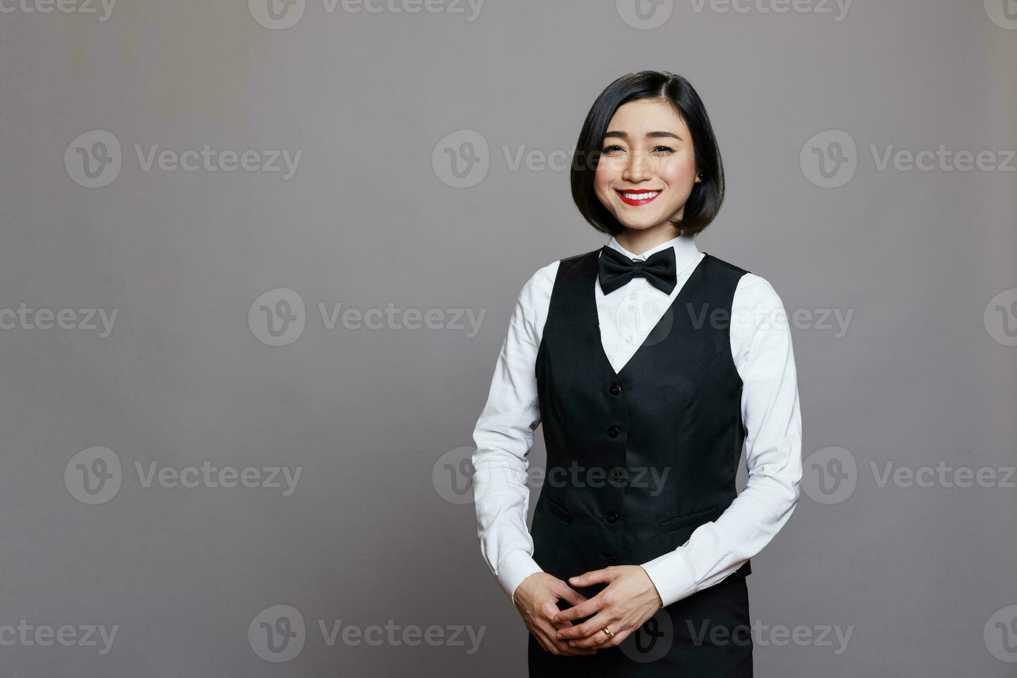 Smiling asian restaurant waitress wearing black and white uniform standing and looking at camera. Cheerful young woman professional receptionist posing for studio portrait photo