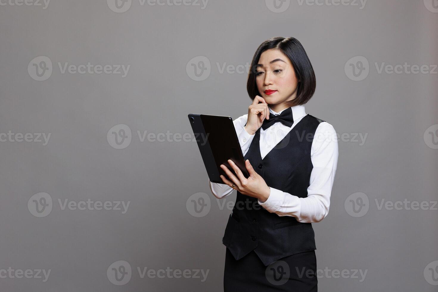 Thoughtful asian waitress in professional uniform managing order on digital tablet. Pensive receptionist thinking while examining restaurant menu on portable gadget and posing in studio photo