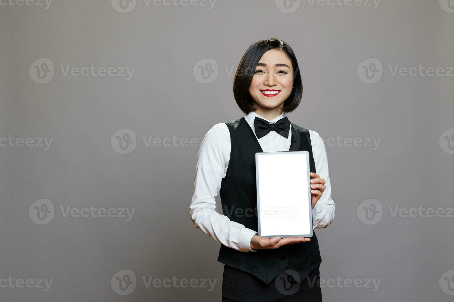 Smiling asian waitress presenting digital tablet empty screen with copy space for advertisement. Friendly restaurant receptionist showing blank touch screen and looking at camera photo