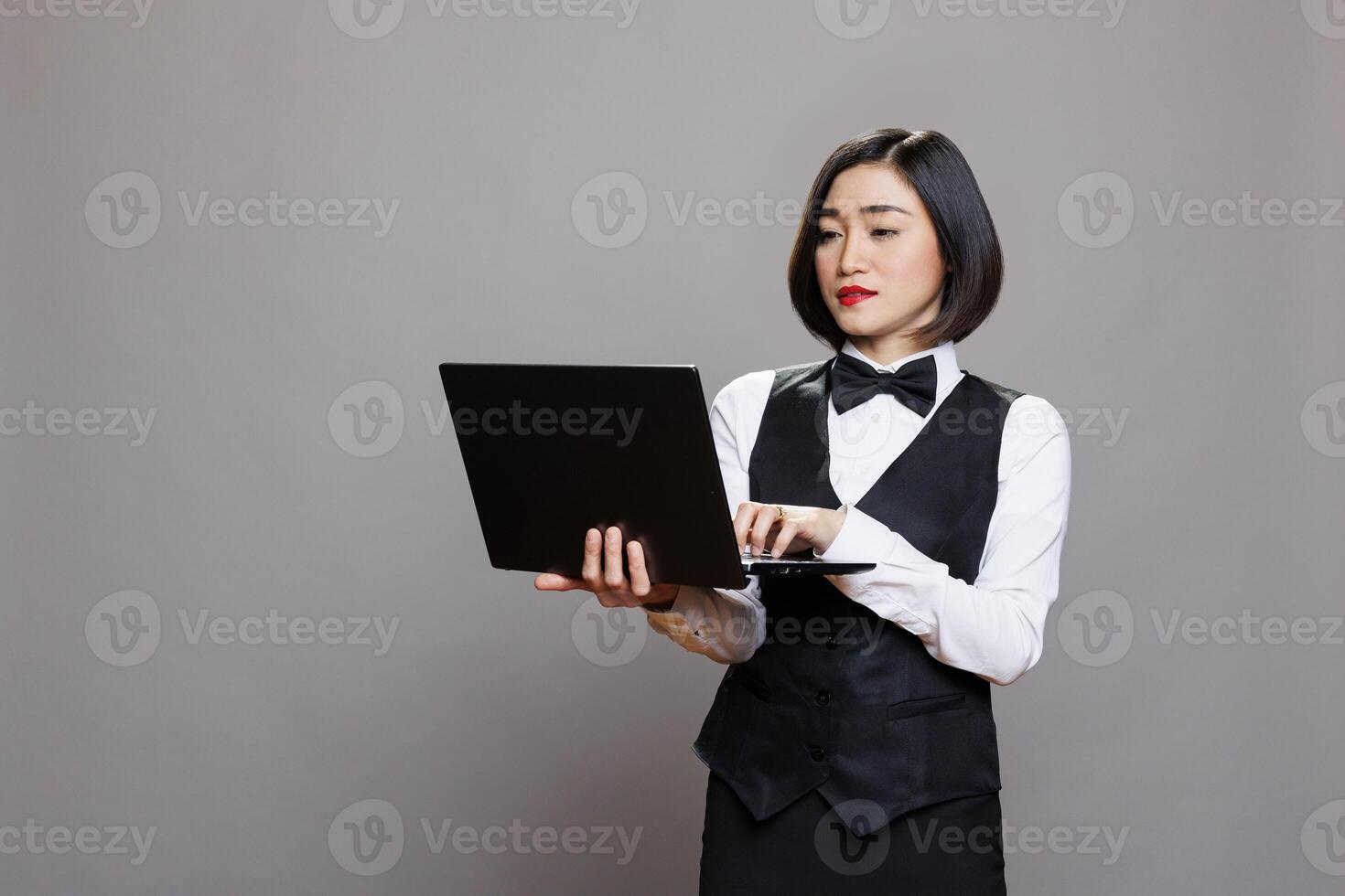 Hospitality service receptionist in uniform working on laptop and typing on keyboard in studio. Young attractive asian waitress holding portable computer and managing orders online photo