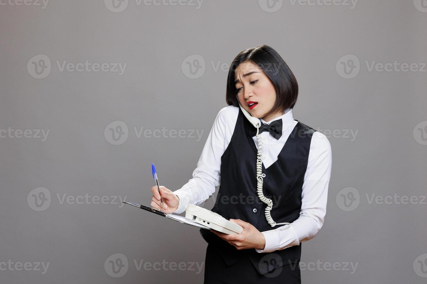 Asian woman receptionist checking information on clipboard while talking with customer on landline phone. Young cafe waitress in uniform taking notes during telephone conversation photo