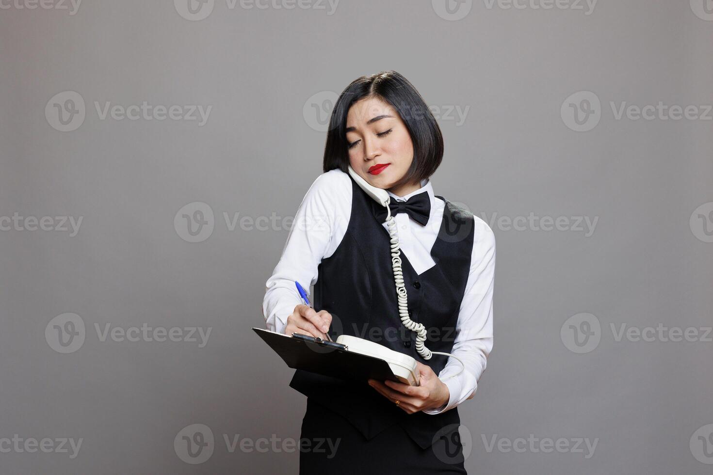 Waitress with clipboard talking on landline phone, managing restaurant operations and customer service. Asian receptionist in uniform taking notes and writing checklist while chatting on telephone photo