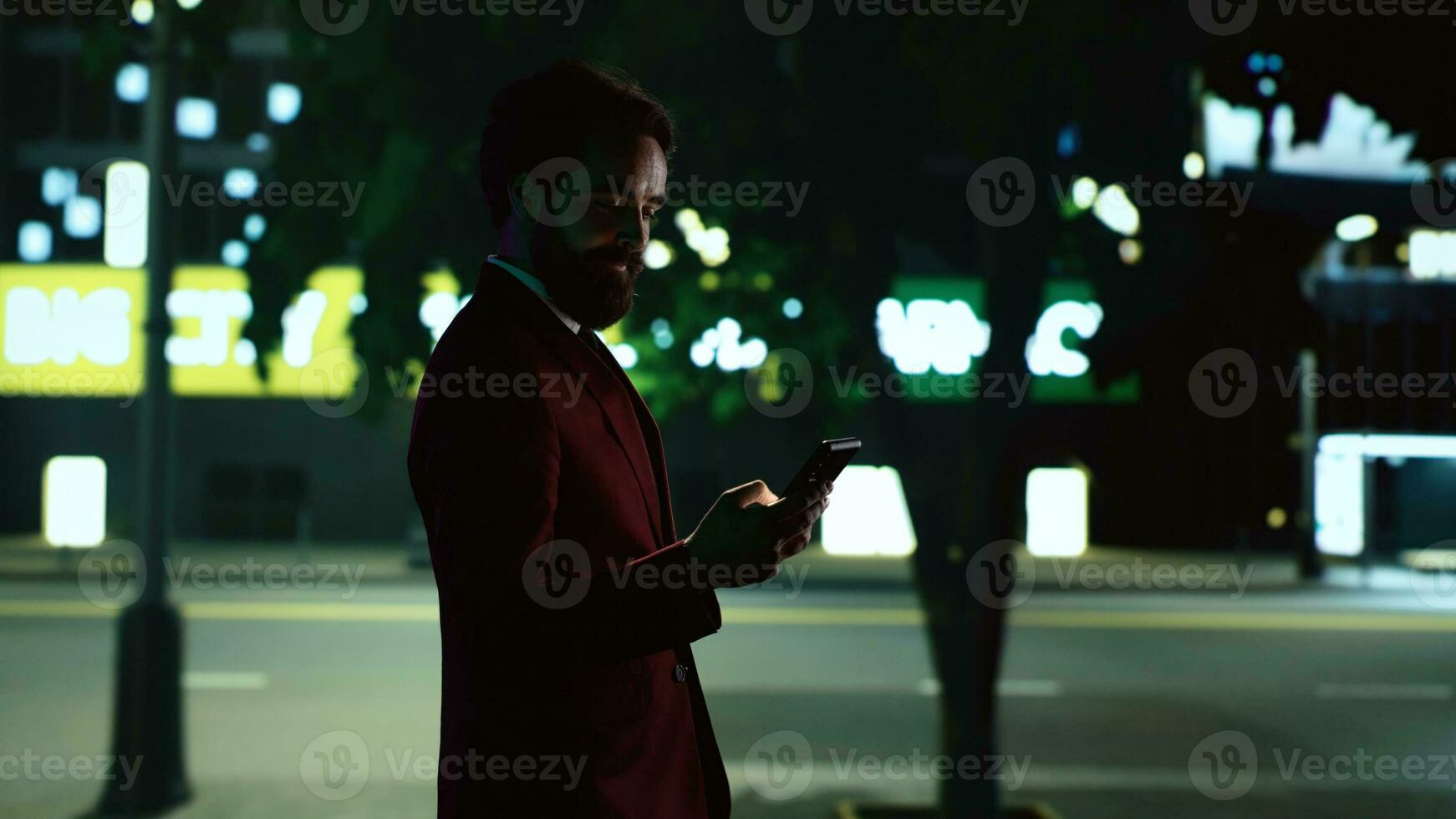 Stylish man walking around town at night, strolling near urban tall building with lights. Entrepreneur walks near skyscrapers in city centre illuminated by streetlamps, texting messages on phone. photo