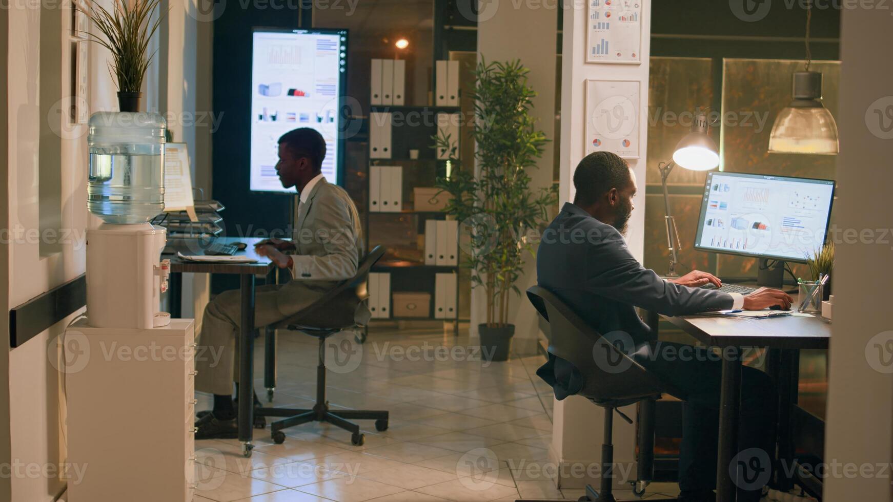 African american employees having fun at work during nightshift, holding race with desk chairs. Relaxed coworking businessmen enjoying overnight stay in office, playing together photo