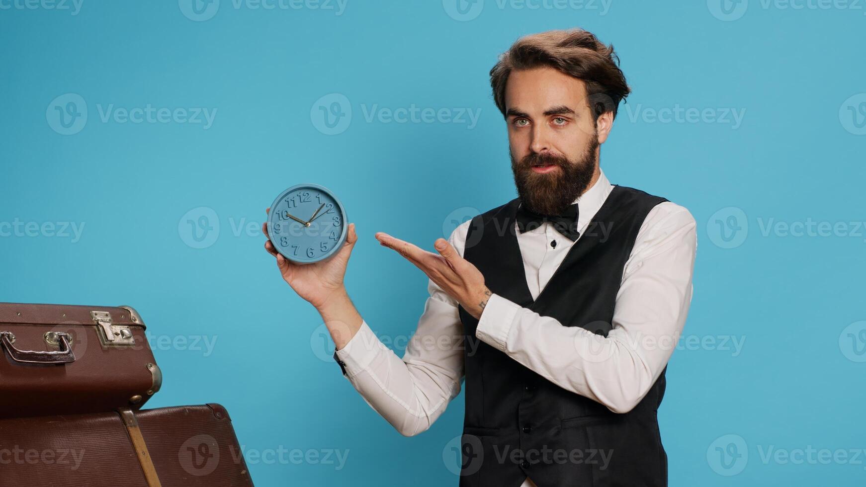 Doorkeeper shows wall clock on camera, checking time and trying to be punctual in hotel travel sector. Man hotel concierge using watch to see hour and minutes in studio next to trolley bags. photo
