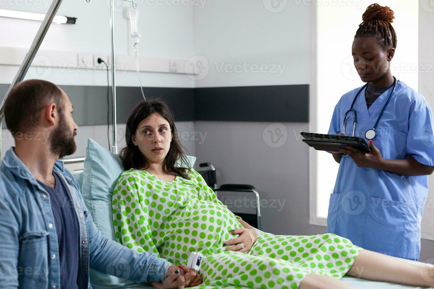 Couple receiving medical assistance from african american nurse before childbirh in hospital ward. Nurse typing woman contractions level on tablet computer, explaining pregnancy labor process photo
