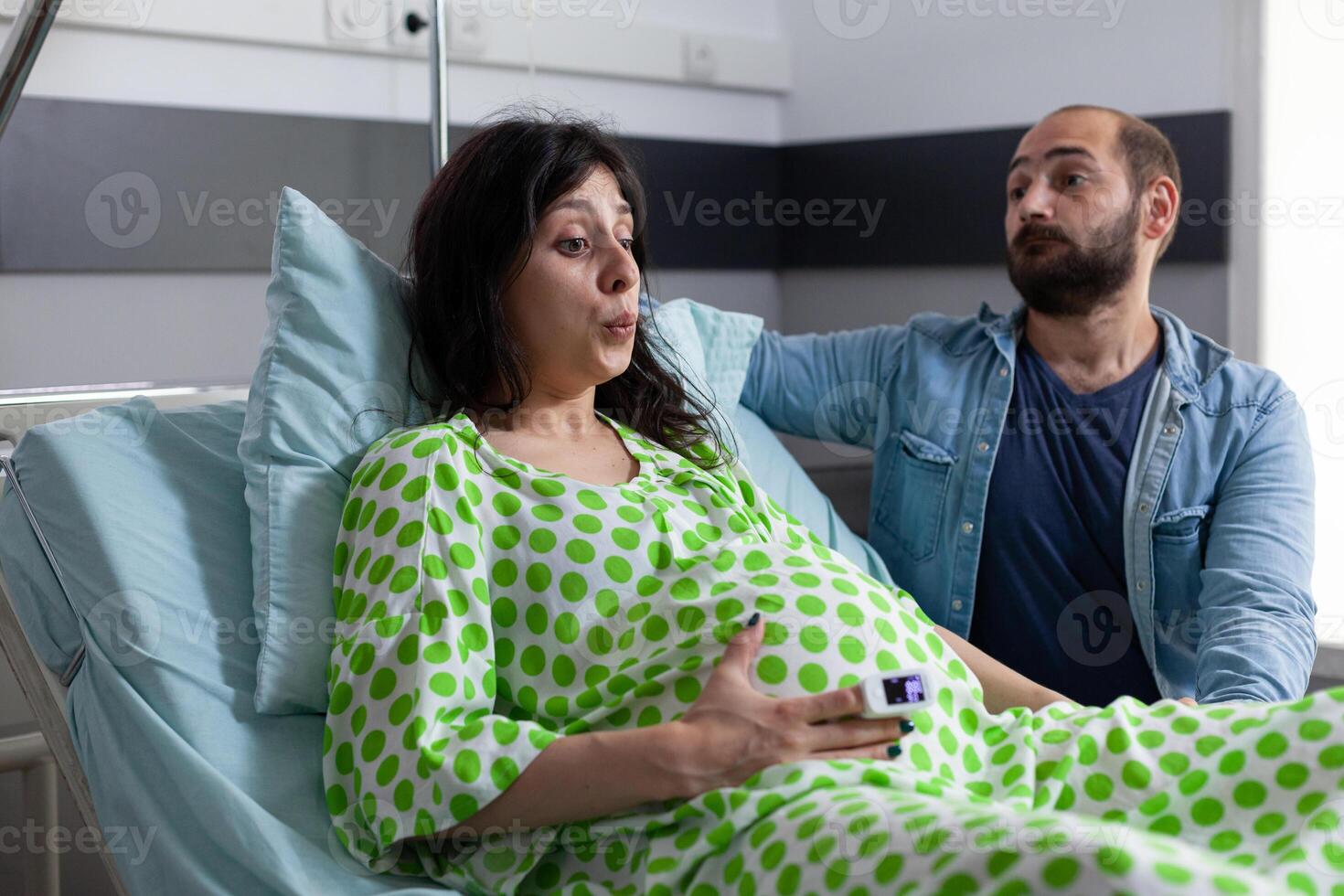 Pregnant woman doing breathing exercices, holding hands on belly while having painful contractions. Patient with pregnancy going into labor in hospital ward, husband standing beside her comforting. photo