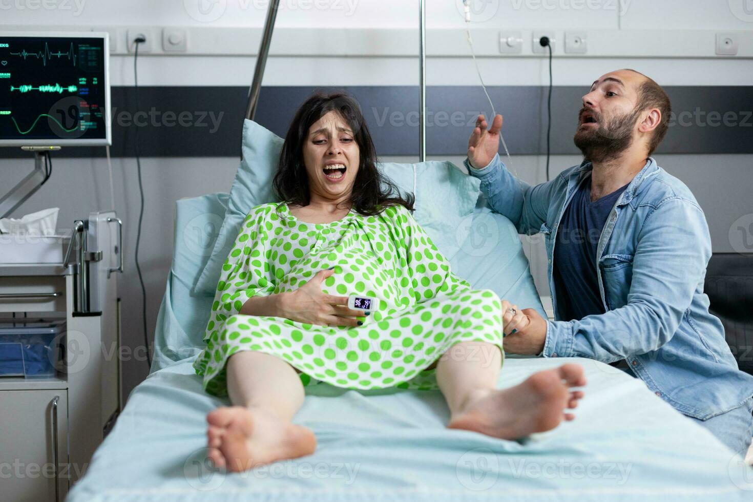 Young couple preparing to be parents in hospital ward bed at healthcare facility. Pregnant woman having painful contractions getting into labor while husband asking for medical assistance photo