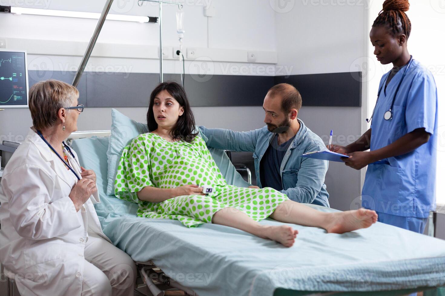 Diverse medical team discussing with pregnant woman before caesarean surgery in hospital ward. Patient with pregnancy lying in bed having contractions, being comforting by husband in maternity clinic photo